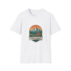 May include: A white short-sleeve t-shirt featuring a central graphic of a mountain landscape with a hiking boot in the foreground. The design is enclosed in a shield-shaped emblem with an orange sky and includes the text TOUCH GRASS on a banner.