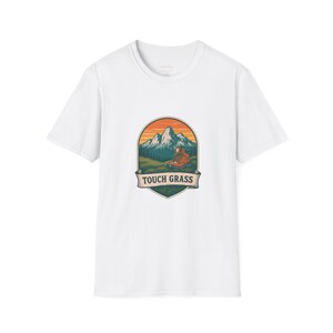 May include: A white short-sleeve t-shirt featuring a central graphic of a mountain landscape with a hiking boot in the foreground. The design is enclosed in a shield-shaped emblem with an orange sky and includes the text TOUCH GRASS on a banner.