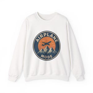 May include: A white long-sleeve sweatshirt featuring a circular graphic on the chest. The design displays a dark blue airplane flying over light blue mountain peaks against an orange sun, with the text AIRPLANE MODE arched around the circle.