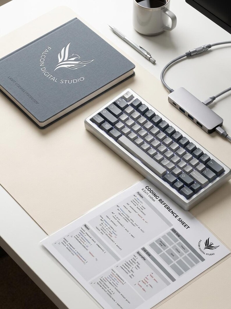 K&ouml;nnte beinhalten: Ein graues Notizbuch mit dem Falcon Digital Studio Logo, eine mechanische Tastatur mit grauen Tasten, ein silberner USB-C-Hub mit Kabeln, ein silberner Stift, eine Kaffeetasse und ein wei&szlig;es Coding-Referenzblatt mit Text und Diagrammen.