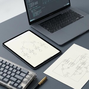 K&ouml;nnte beinhalten: Ein grauer Laptop mit Code-Anzeige, ein Tablet mit einem Flussdiagramm, ein wei&szlig;es Blatt Papier mit einem gedruckten Logikdiagramm, eine mechanische Tastatur mit grauen Tasten und ein schwarz-goldener Stift auf einer flachen Oberfl&auml;che.