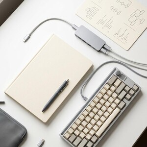 May include: A mechanical keyboard with grey and beige keycaps, a grey USB-C multi-port hub with connected cables, a beige notebook, a grey pen, and a grey zippered pouch. A paper sheet with printed flowcharts is positioned near the hub.