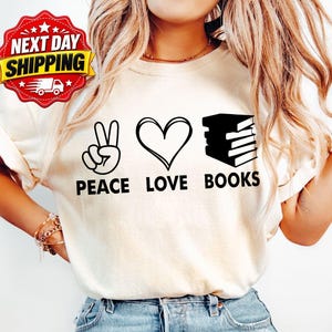 Op de afbeelding: Een crèmekleurig T-shirt met een zwart grafisch ontwerp met een vredesteken, een hart en een stapel boeken. Onder de iconen staat de tekst PEACE LOVE BOOKS in een vetgedrukt, zwart schreefloos lettertype. Het T-shirt heeft korte mouwen.