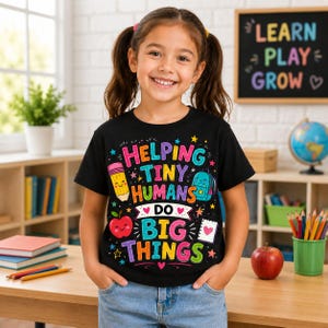 May include: A black short-sleeved t-shirt featuring a colorful graphic with the text "HELPING TINY HUMANS DO BIG THINGS" surrounded by illustrations of a smiling pencil, a backpack, an apple, a notebook, and various stars and hearts.