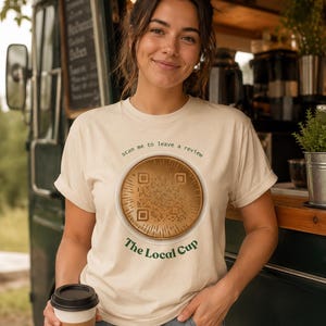 Könnte beinhalten: Ein cremefarbenes Kurzarm-T-Shirt mit einer zentralen Grafik eines goldfarbenen, kreisförmigen QR-Codes. Über der Grafik steht der Text "scan me to leave a review" und darunter der grüne Schriftzug "The Local Cup" in einer Serifenschrift.