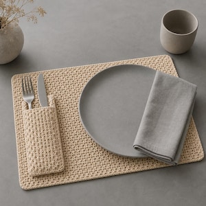 May include: A rectangular beige crocheted placemat featuring an integrated pocket for cutlery. The set includes a silver fork and knife tucked into the pocket, a round matte grey plate, and a folded grey linen napkin placed on the placemat.