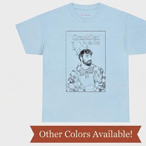 Puede incluir: Una camiseta de manga corta color azul claro con un gráfico de líneas en blanco y negro de una persona con barba que lleva un sombrero alto a rayas y una pajarita. El texto sobre la figura dice "CrankDat in da Hat".