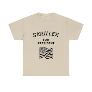 May include: A beige short-sleeve t-shirt featuring the black text SKRILLEX FOR PRESIDENT printed in a bold font. Below the text is a graphic illustration of a waving American flag rendered in black and white line art on the centre chest.