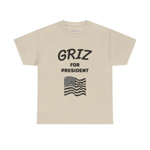 Op de afbeelding: Een beige t-shirt met korte mouwen en de zwarte tekst GRIZ FOR PRESIDENT in het midden gedrukt. Onder de tekst staat een grafische illustratie van een wapperende Amerikaanse vlag in zwarte inkt. Het shirt heeft een ronde hals en pasvorm.