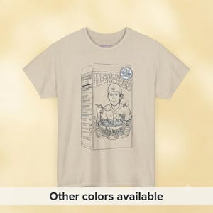 May include: A beige short-sleeve t-shirt featuring a black line art graphic of a cereal box labeled "ILLENI-YUMS". The box depicts a person wearing a baseball cap eating from a bowl with a spoon, with a circular badge reading "THE NATURE KING".