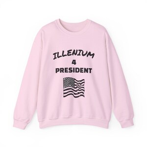 May include: A light pink long-sleeve sweatshirt featuring the black text ILLENIUM 4 PRESIDENT printed in the center. Below the text is a black graphic of a waving American flag with a star-filled canton and horizontal stripes.