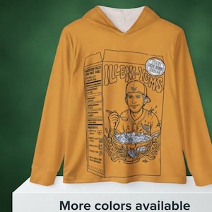 May include: A mustard yellow long-sleeve hooded sweatshirt featuring a large graphic print of a cereal box labeled Illeni-Yums. The graphic depicts a person wearing a baseball cap eating from a bowl with a spoon, alongside nutrition facts.