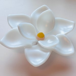 Pode incluir: Um objeto em forma de flor branca com pétalas lisas e curvas dispostas em camadas. O centro da flor apresenta um pequeno detalhe circular texturizado em amarelo-alaranjado. A aparência geral é brilhante e sólida, como uma flor esculpida.
