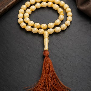 May include: A prayer bead strand featuring smooth, pale yellow, oval-shaped beads arranged in a circular loop. The strand includes a central elongated bead piece that connects to a long, dark brown, fine-threaded tassel hanging downwards.