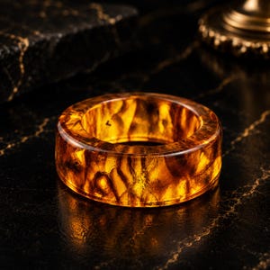 Op de afbeelding: Een brede, ronde ring gemaakt van doorschijnend, barnsteenkleurig materiaal. Het oppervlak heeft een wervelend, gemarmerd patroon van diepe gouden, oranje en donkerbruine tinten die het licht vangen, wat zorgt voor een gloeiende, semi-transparante uitstraling.