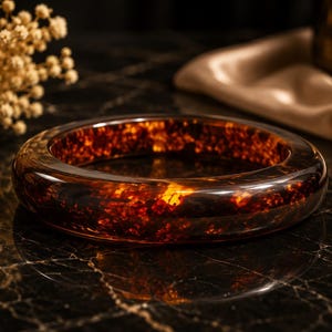 May include: A circular bangle bracelet crafted from translucent amber-colored resin, featuring a polished, glossy finish with internal golden and dark brown flecks that create a mottled, light-catching appearance throughout the thick, rounded band.