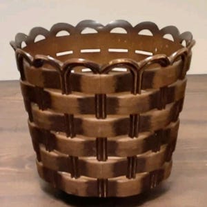 Peut inclure: Un panier rond marron avec un motif tressé et un bord supérieur festonné. La structure présente des bandes verticales et horizontales créant un aspect de grille, avec un design découpé décoratif le long du bord supérieur du récipient.