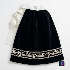 Puede incluir: Una falda de terciopelo negro con una cintura ancha blanca y lazos laterales largos. El dobladillo está decorado con una banda horizontal de intrincado bordado de encaje blanco en un patrón festoneado repetitivo, contrastando con la tela oscura.