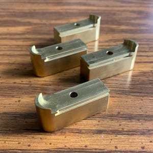 May include: Four rectangular brass components with a metallic gold finish. Each piece features a rounded end, a flat top surface with a centered circular hole, and a recessed channel running along the length of the top edge of the block.