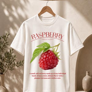 Puede incluir: Una camiseta blanca de manga corta con un gráfico de una frambuesa roja con hojas verdes. El texto RASPBERRY HARVEST SEASON aparece en la parte superior, junto a un párrafo descriptivo sobre el sabor y la estructura de la baya en rojo.