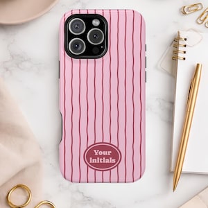 Puede incluir: Una funda para smartphone de color rosa claro con un patrón de líneas verticales onduladas de color rosa oscuro. En la parte inferior central hay un emblema ovalado rosa oscuro con el texto "Your initials" en una fuente serif rosa claro.