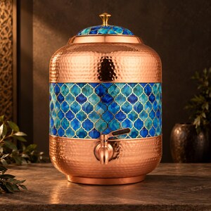 Puede incluir: Un dispensador de agua de cobre martillado con una banda central de patrón de cuatrifolio en tonos azul y turquesa. El recipiente incluye una tapa a juego con un pomo dorado y un grifo color cobre con una palanca negra.