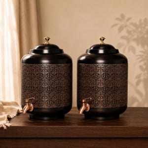 May include: Two cylindrical black beverage dispensers featuring a detailed copper-colored damask pattern around the center. Each dispenser has a matching black lid topped with a small gold-toned finial and a copper-colored spigot at the base.