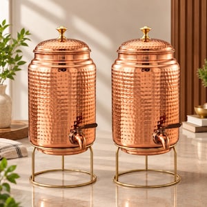 May include: Two cylindrical copper beverage dispensers with a hammered texture, each featuring a gold-toned lid with a knob handle and a matching gold-toned metal wire stand. A small metal spigot is attached to the lower front of each vessel.