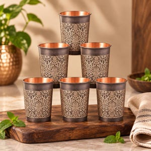 Könnte beinhalten: Ein Set aus sechs zylindrischen Metallbechern, die in einer Pyramidenform angeordnet sind. Jeder Becher hat eine dunkle, verwitterte Außenfläche mit einem komplizierten, hellen floralen Damastmuster und einem glatten, kupferfarbenen Rand.