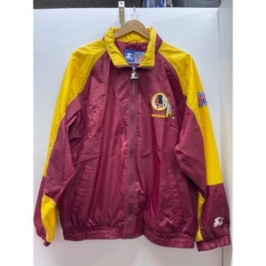 May include: A burgundy and yellow zip-up windbreaker jacket featuring the Washington Redskins logo on the chest, an NFL patch on the sleeve, and a Starter brand logo on the cuff and zipper pull. The jacket has a high collar and long sleeves.