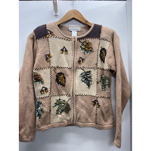 Op de afbeelding: Een beige gebreid vest met rits en een patchworkontwerp met afwisselende lichtbeige en lichtbruine vierkanten. Het kledingstuk is versierd met geborduurde herfstbladeren en eikels in tinten groen, bruin en geel, met donkerbruine schouderstukken.
