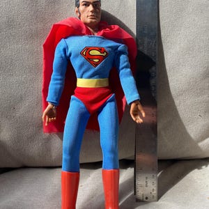 May include: A Superman action figure wearing a blue fabric suit with a red cape, red trunks, a yellow belt, and red boots. The figure features a red and yellow chest emblem. A metal ruler placed next to the figure shows a height of 12 inches.