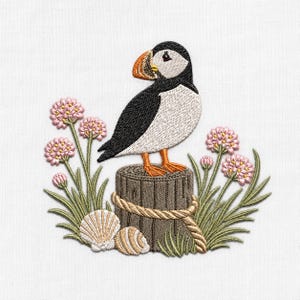 May include: An embroidery design featuring a puffin with black, white, and orange plumage perched on a brown wooden post wrapped in rope. The scene includes light pink flower clusters on green stems and two white seashells at the base.