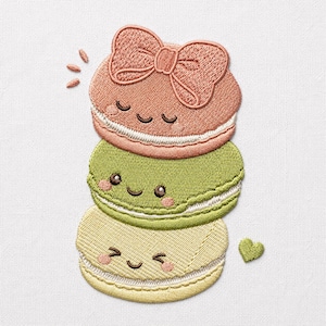 Puede incluir: Un parche bordado que muestra una pila de tres macarons coloridos con caras sonrientes. El macaron superior es de color melocotón con un lazo, el del medio es verde claro y el inferior es amarillo pálido. Cada uno tiene relleno blanco.