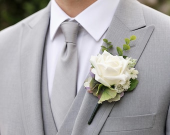 May include: A light grey three-piece suit with a white collared shirt and a matching grey necktie. A floral buttonhole is pinned to the lapel, featuring a single white rose, small green eucalyptus leaves, and tiny white filler flowers.