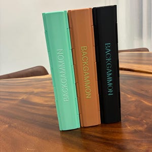 May include: Three rectangular backgammon game cases standing upright side by side. The left case is mint green, the middle case is terracotta orange, and the right case is black. Each case features the word BACKGAMMON printed vertically in capital letters.