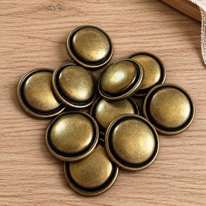 May include: A collection of round, convex buttons featuring a metallic antique brass finish. Each button has a smooth, domed center surrounded by a thin, raised circular rim. The buttons have a weathered, darkened patina in the recessed areas.