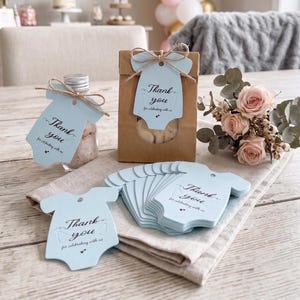 May include: Light blue paper tags shaped like baby onesies with the printed text Thank you for celebrating with us. The tags feature a small hole at the top and are tied with brown twine to a glass bottle and a brown paper bag containing treats.