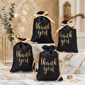 May include: Four black satin drawstring gift bags featuring the gold text thank you printed on the front. Each bag is tied with a gold ribbon. The bags are arranged on a white marble surface with gold veining, alongside gold jewelry pieces.