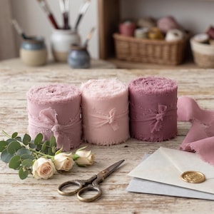 May include: Three spools of frayed-edge fabric ribbon in varying shades of dusty pink and mauve, tied with matching ribbon scraps. A small sprig of eucalyptus with two cream-colored roses, a pair of metal scissors, and a cream envelope with a gold wax seal.