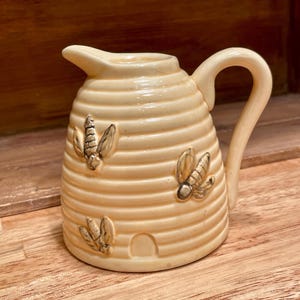 May include: A cream-colored ceramic creamer shaped like a beehive, featuring horizontal ridges, a curved handle, a small pouring spout, and three raised bee motifs with a small arched entrance at the base of the pitcher.