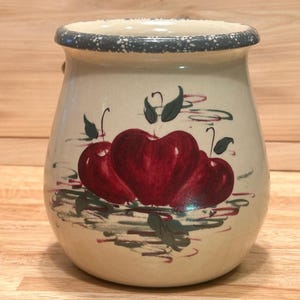May include: A cream-colored ceramic crock featuring a hand-painted design of three red apples with green leaves. The vessel has a rounded body and a wide opening with a dark grey speckled rim. The painted apples are centered on the front surface.