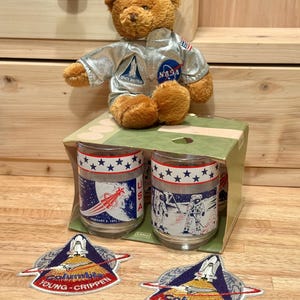 May include: A brown plush teddy bear wearing a silver NASA space suit, sitting on a cardboard box containing two glass tumblers with space-themed graphics. Two embroidered patches featuring a space shuttle and the text Columbia Young Crippen.