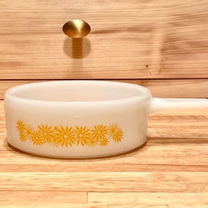 May include: A white milk glass saucepan featuring a horizontal band of yellow daisy flowers around the exterior. The round vessel has a single integrated handle extending from one side and a smooth, opaque finish throughout the entire piece.