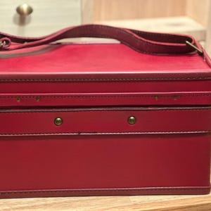 May include: A rectangular red storage case with a flat top and a single handle attached by metal hardware. The exterior features stitched edges and two small brass-colored studs on the front panel, providing a structured and functional design.