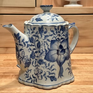May include: A white ceramic teapot featuring a blue floral pattern with intricate vines and leaves. The teapot has a scalloped base and rim, a curved spout, a handle, and a matching lid with a small knob handle on top, all decorated in blue.