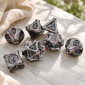 May include: A set of seven polyhedral dice featuring a metallic silver finish with dark recessed detailing and scattered red paint accents. Each die has a complex, textured surface with raised numbers, including a d20, d12, d10, d8, d6, d4, and a d100.