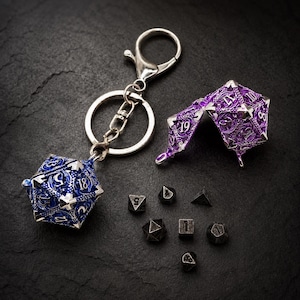 May include: A blue metal twenty-sided die keychain with intricate silver filigree patterns and white numbers, a matching purple openable twenty-sided die container, and six small black metal polyhedral dice scattered on a dark textured surface.