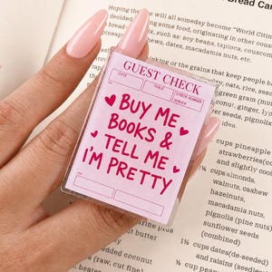 May include: A rectangular clear acrylic keychain featuring a light pink guest check design with dark pink text that reads GUEST CHECK, BUY ME BOOKS & TELL ME I'M PRETTY, accented with small heart icons and blank fields for date, table, and server.