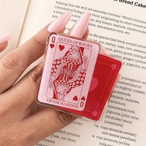 Può includere: Un ciondolo quadrato in acrilico con il design di una carta da gioco Regina di Cuori e la scritta Queen of Books. L'illustrazione raffigura una figura stilizzata con occhiali a forma di cuore che tiene un fiore, nei toni del rosa e del rosso.